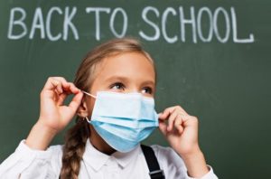 young girl with a face mask in front of a blackboard reading back to school