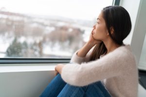 Sad looking youg woman sits at a window gazing at a grey winter landscape. She may be experiencing seasonal affective disorder (SAD)