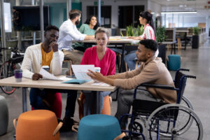 professional people sitting at tables in a cafeteria. In the front table, there are two young black men and a young white woman. One of the men is in a wheelchair. They are sharing and discussing papers and catching up on HR news.