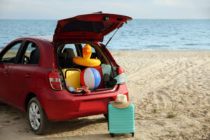 red car on a beach in the summer. The trunk is open, displayinf colorful luggage and summer topys like a beachball and a duck shaped floatie, Image to illustrate blog post on summer safety.
